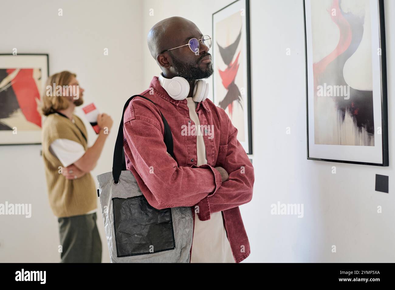 Mann mit Kopfhörern, der große abstrakte Kunst an der Galeriewand untersucht, während eine andere Person im Hintergrund mit Kunst beschäftigt, die sich auf ein anderes Stück konzentriert Stockfoto