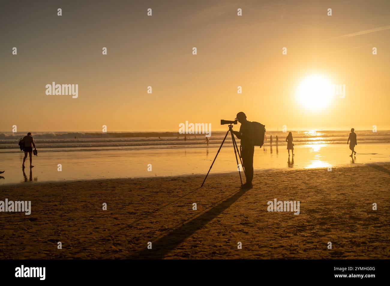 Fotograf in voller Silhouette steht am Stativ mit Kamera und langem Objektiv am Strand bei Sonnenuntergang. Eine ferne Figur im goldenen Licht fängt einen ruhigen Abend ein, bei dem Menschen entlang der Küste spazieren Stockfoto