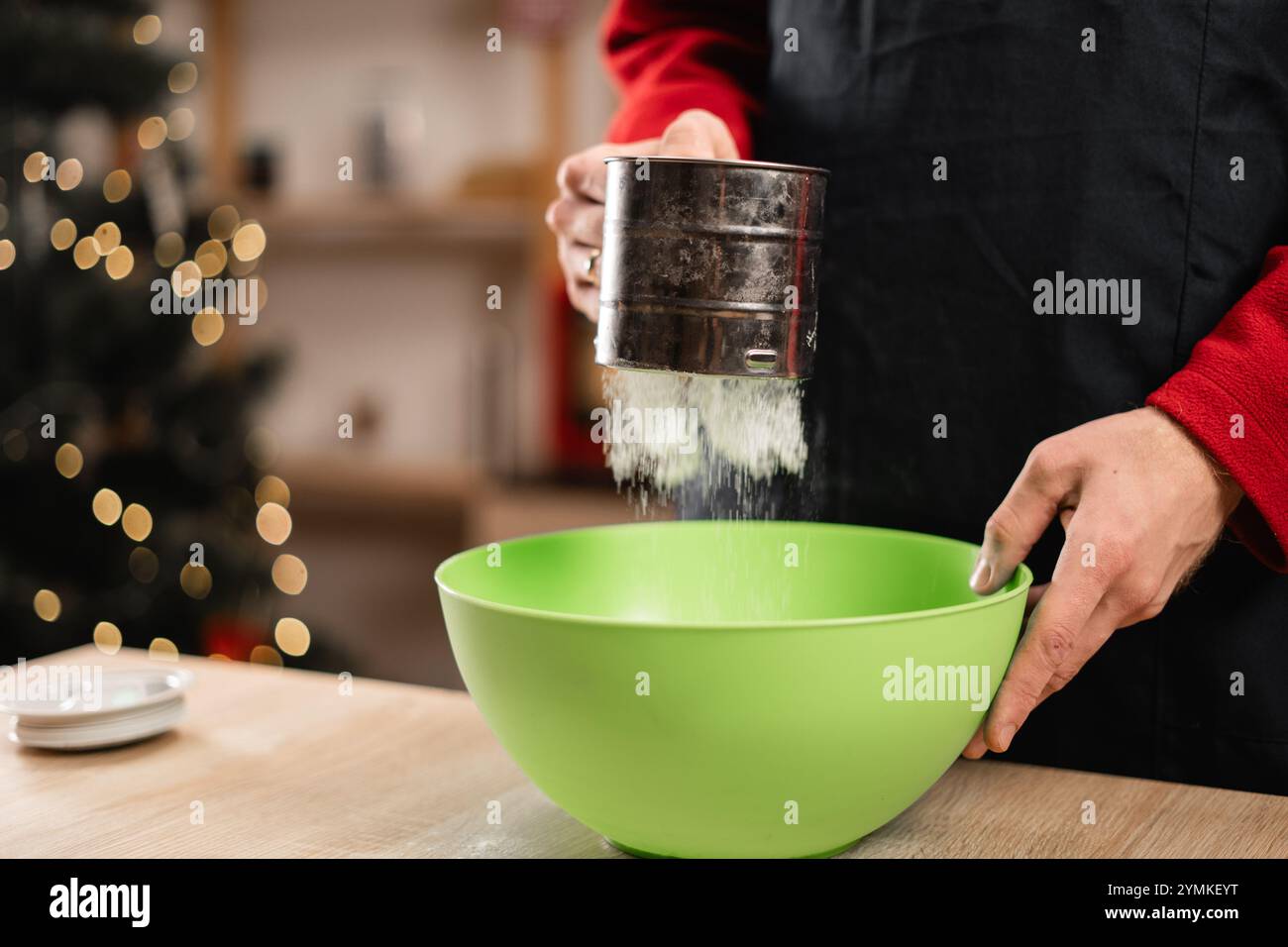 Mann siebt Mehl durch Sieb in eine Schüssel, während er zu Hause an Weihnachten backt. Mehlstaub sichtbar in der Luft, dynamische Szene. Backzutaten Stockfoto