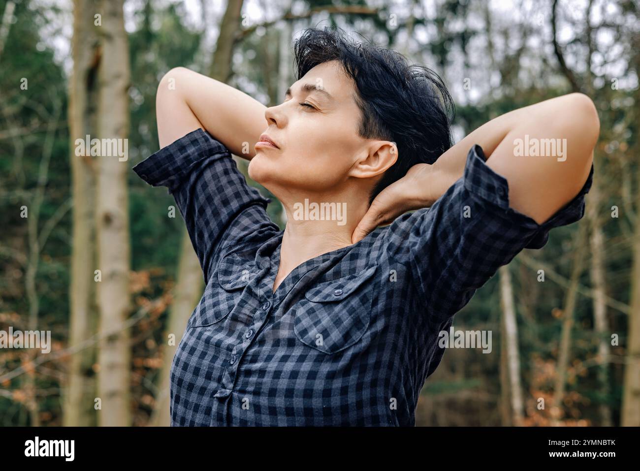 Glückliche Frau, 45-50 Jahre alt, trägt ein kariertes Hemd, genießt die frische Luft im Wald mit ihren Händen hinter ihrem Kopf Stockfoto