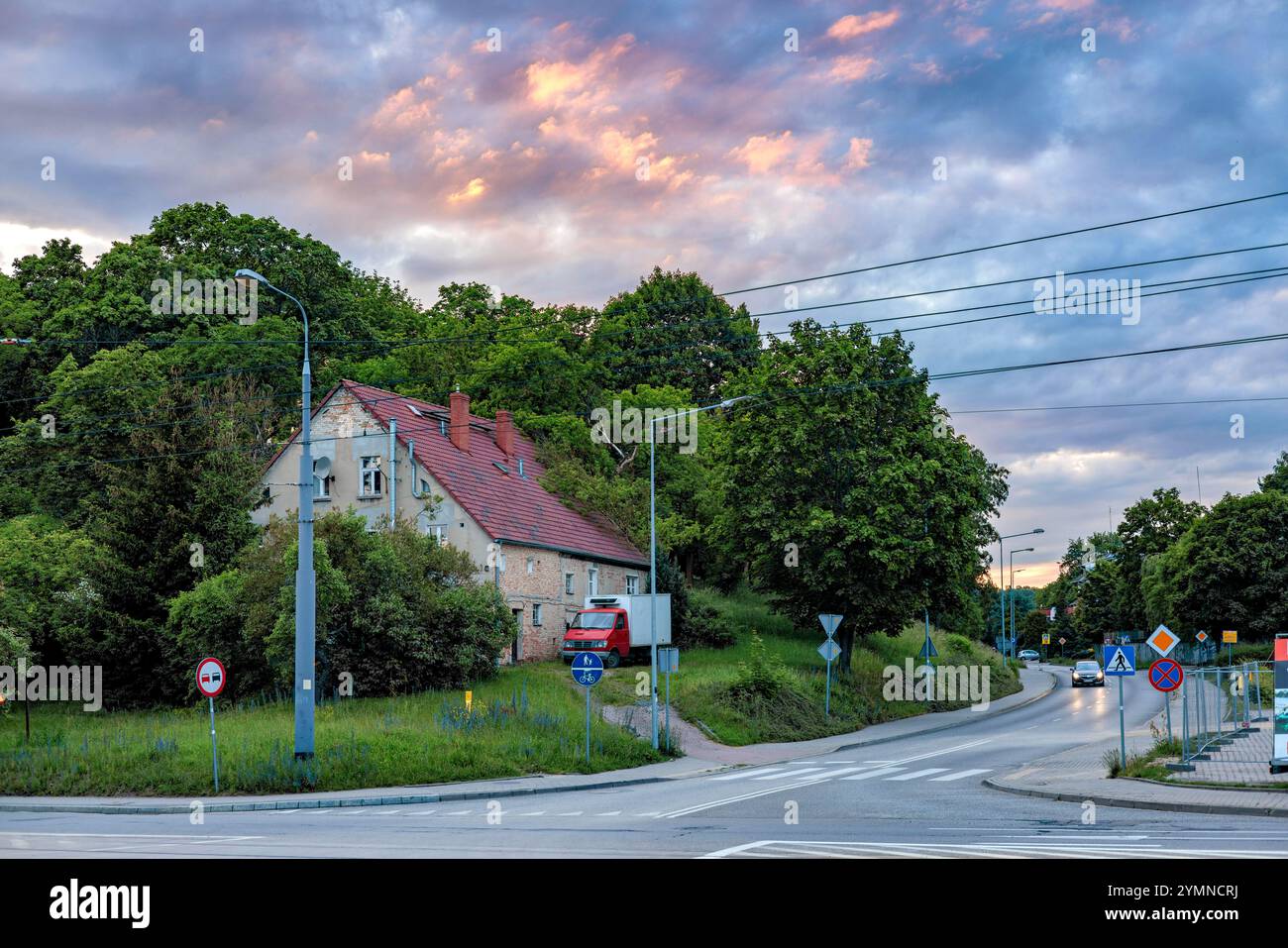 Ein Haus mit einem roten Ziegeldach, das zwischen den Bäumen eines Stadtparks vor dem Hintergrund eines wunderschönen Sonnenuntergangs steht. Kreuzungen mit Straßenschildern Stockfoto