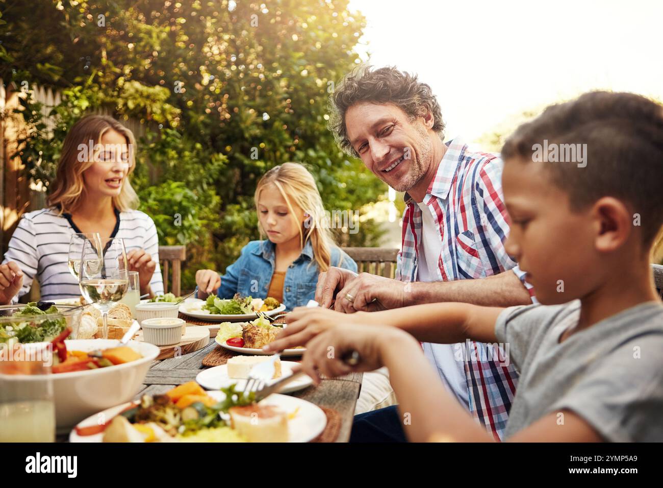 Mom, Dad und Kinder beim Mittagessen im Freien mit einem Lächeln, Feiern und fröhlichen Familienessen im Hinterhof. Vater, Mutter und Kinder am Tisch Stockfoto