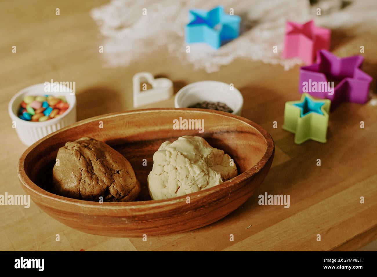 Eine Holzschale mit Keksteig, verschiedene Formen zum Schneiden und bunte Süßigkeiten auf dem Tisch, die eine ideale Einrichtung für hausgemachte Backaktivitäten schaffen Stockfoto