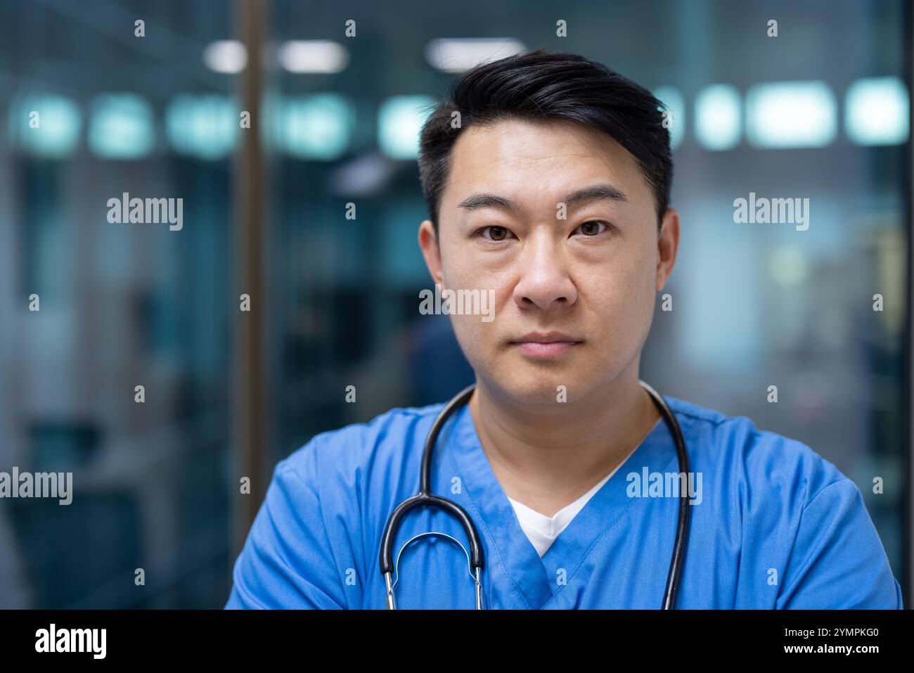 Asiatischer Mann Arzt in blauen Peelings steht selbstbewusst mit Stethoskop in moderner Gesundheitseinrichtung. Ausdruck ernst, Projizierung von Professionalität und Hingabe im medizinischen Bereich. Stockfoto