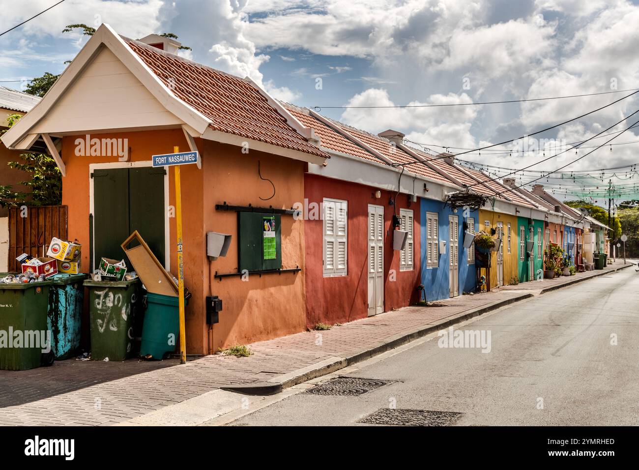 Eine Reihe kleiner, einstöckiger Häuser aus der Kolonialzeit in Terrakotta, Rot, Blau und Grün am Fort Nassauweg in Willemstad, Curacao. Diese einfachen Arbeiterhäuser stehen im Kontrast zu den raffinierteren historizistischen Fassaden, die in anderen Teilen der Stadt zu finden sind. Die Lichter deuten darauf hin, dass die Straße für Festivals oder Märkte genutzt wird. Farbenfrohe Fassaden an renovierten Häusern in der Bargestraat zwischen Schrloo und Fleur de Marie, die mit hellen Dekorationen bedeckt ist, Kòrsou Stockfoto