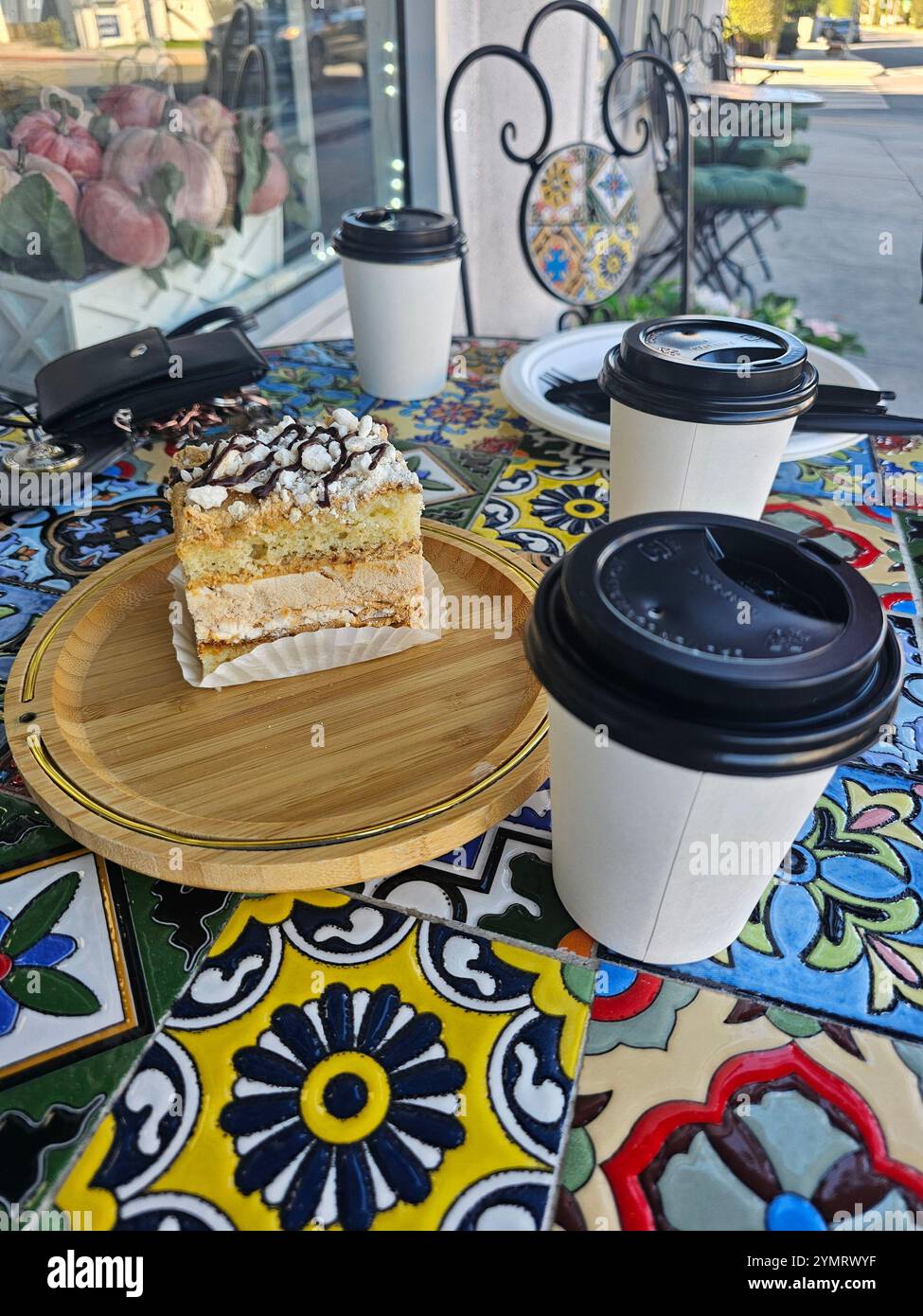 Künstlerisch gestalteter Kaffeetisch im Freien mit bunten Fliesen, einem köstlichen mehrschichtigen Kuchen auf einem Holzteller und Kaffeetassen zum Mitnehmen. Stockfoto