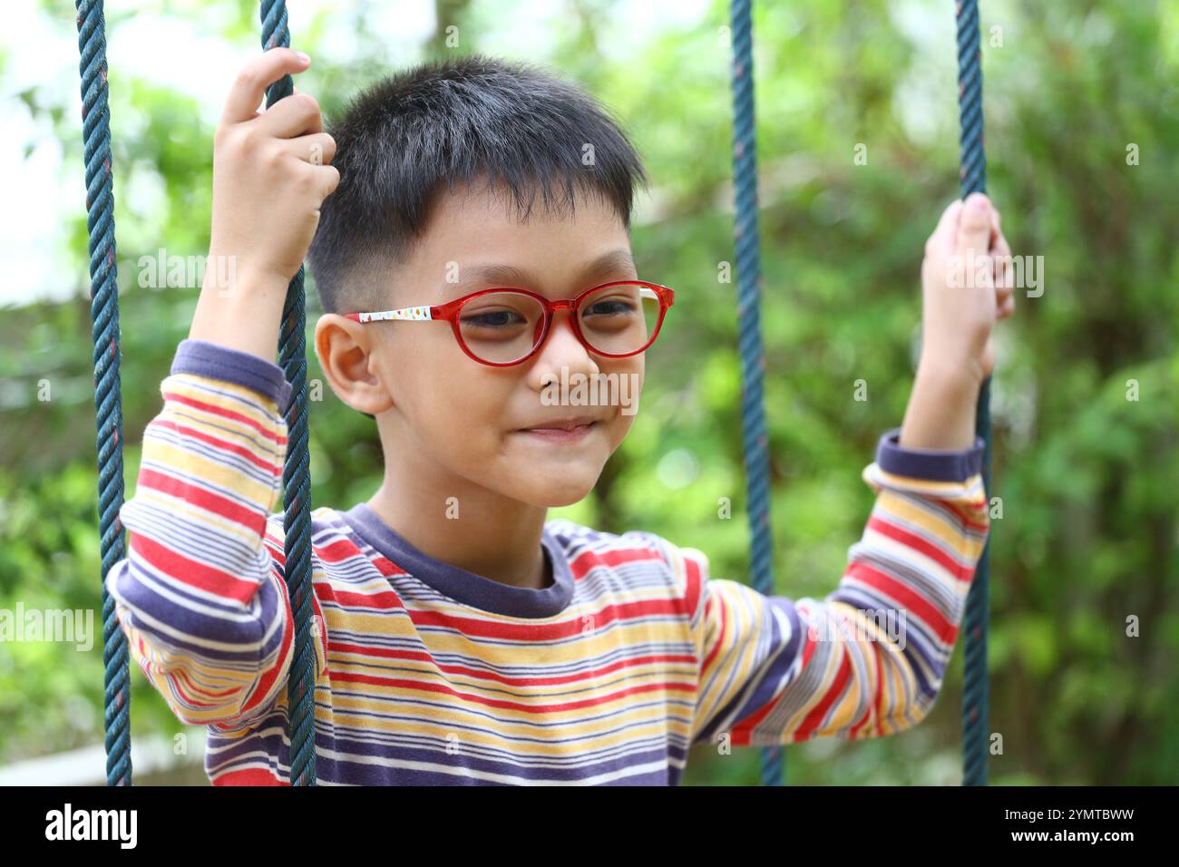 Ein charmanter Junge mit Brille schwingt gern in einem üppigen Garten. Sein spielerischer Ausdruck fängt das Wesen der Kindheit und unbeschwerten Momente ein. Stockfoto