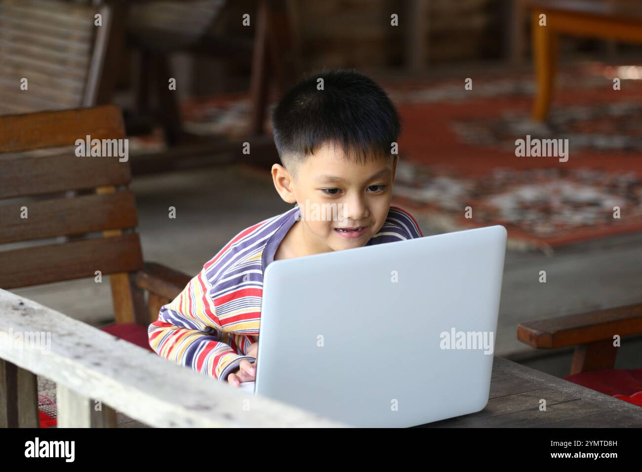 Ein fröhliches Kind sitzt auf Holzmöbeln, in einem Laptop. Die Umgebung ist gemütlich und zeigt die Freude am Lernen und Spielen durch Technologie. Stockfoto