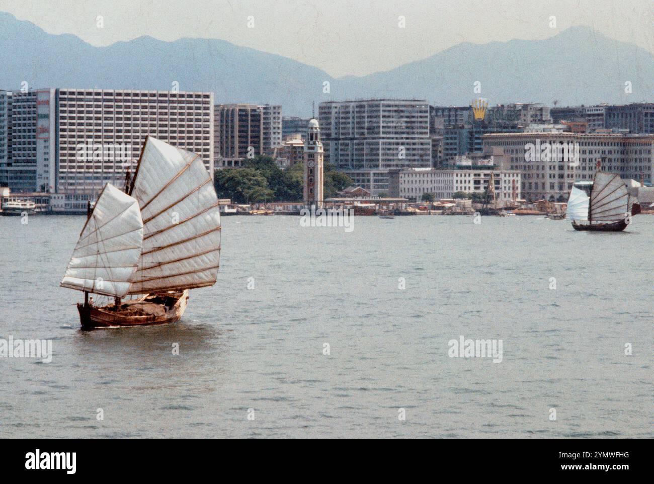 Zwei traditionelle chinesische Fracht-Dschunken, die durch die enge zwischen Hong Kong Island und Kowloon fahren, allein unter Segelkraft. Schon damals (1970er) waren Segel-Junks ein seltener Anblick, besonders in den Narrows, dem geschäftigsten Teil des Hafens. Star Ferry, Kowloon Tower und Peninsula Hotel, Kowloon Side, im Hintergrund. Stockfoto
