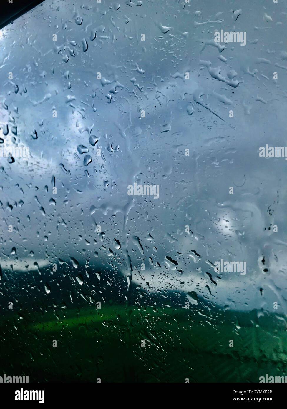 Regentropfen streuen sich über das Glas und schaffen einen ruhigen Moment, während die Grün- und Grautöne der Natur nahtlos im Hintergrund verschmelzen Stockfoto