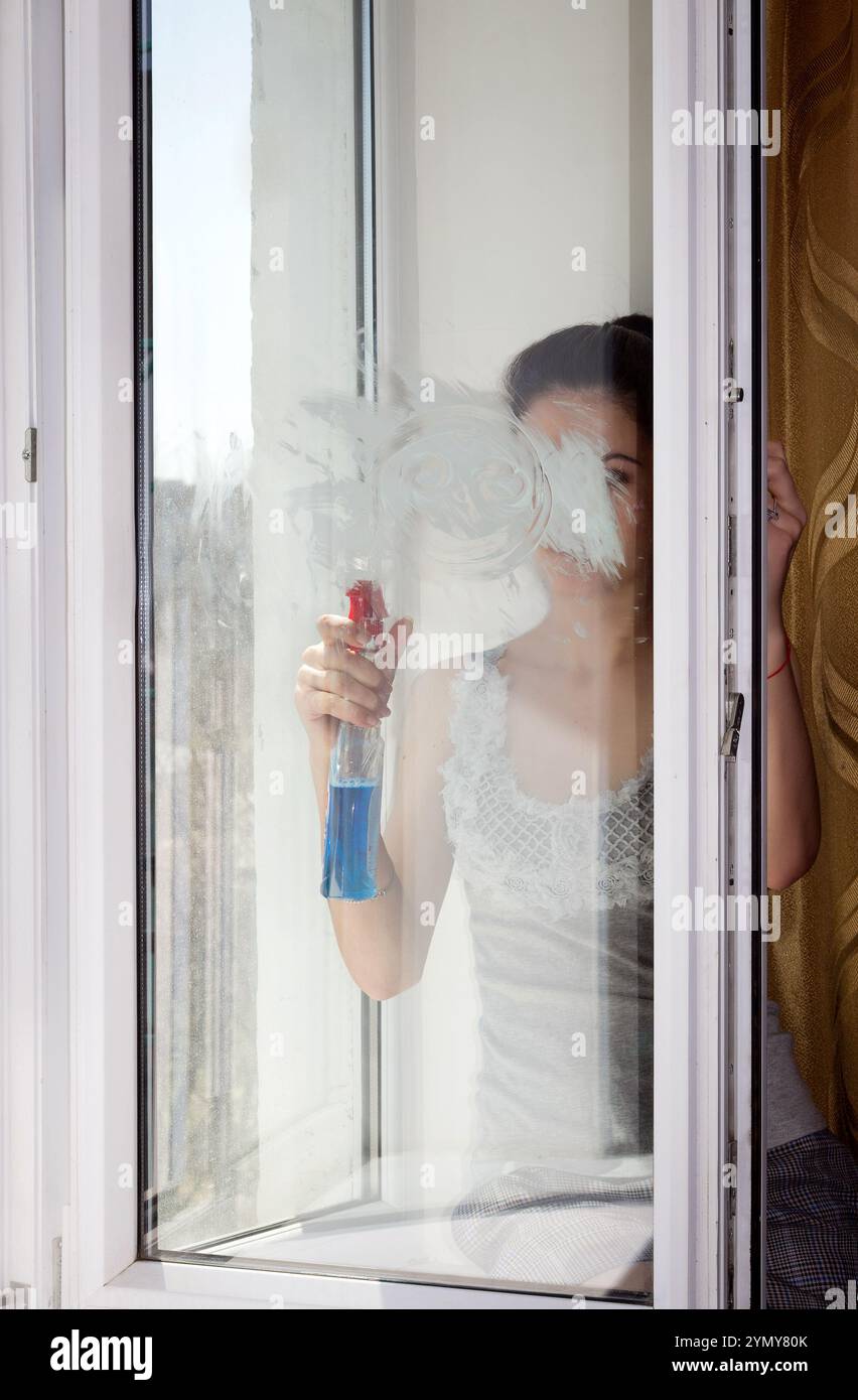 Das junge Mädchen sitzt auf einer Fensterbank und wäscht und reinigt ein Fenster sorgfältig Stockfoto