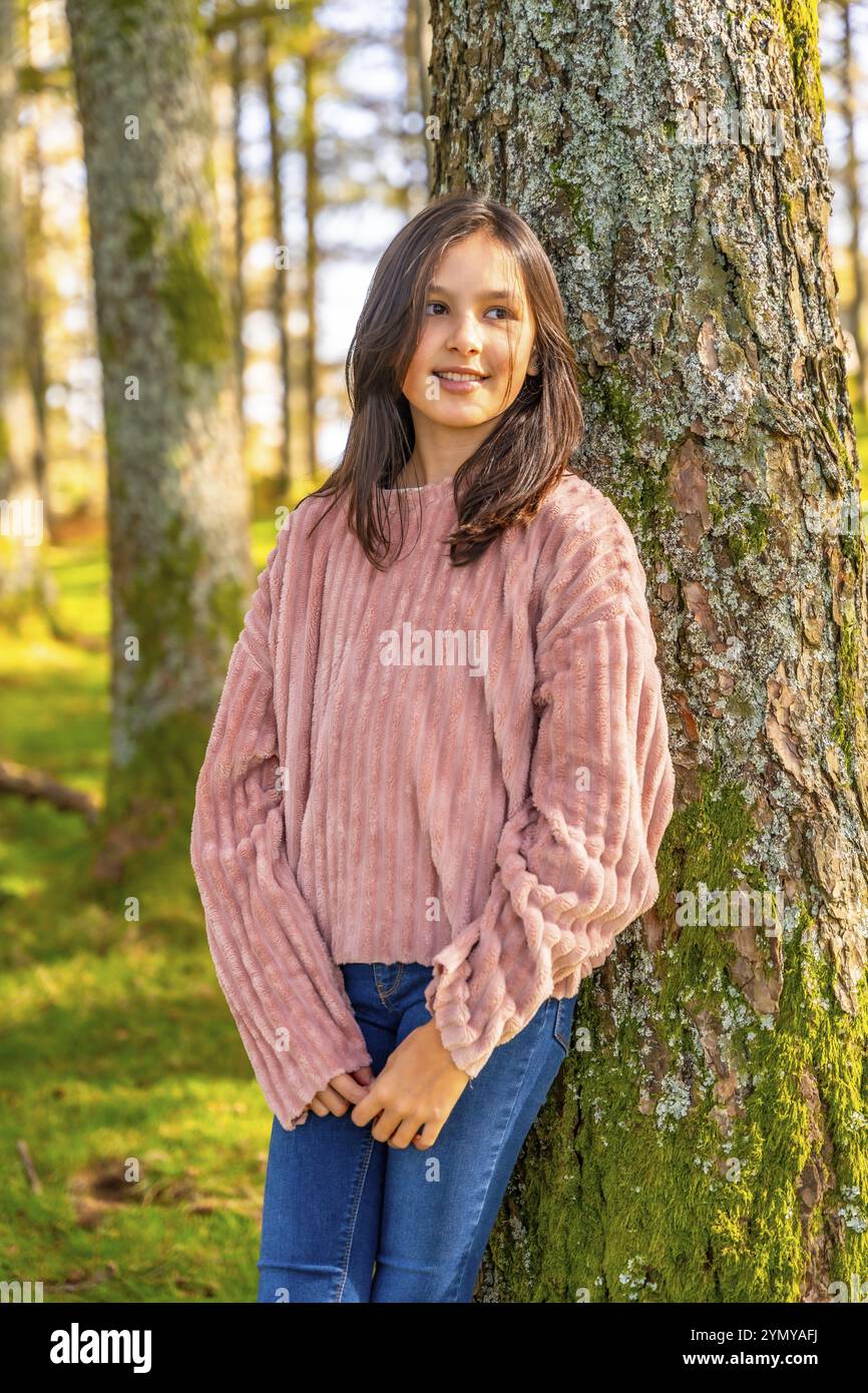 Vertikales, dreiviertel langes Foto eines kaukasischen Mädchens, das sich entspannt auf einen Baumstamm im Wald lehnt Stockfoto