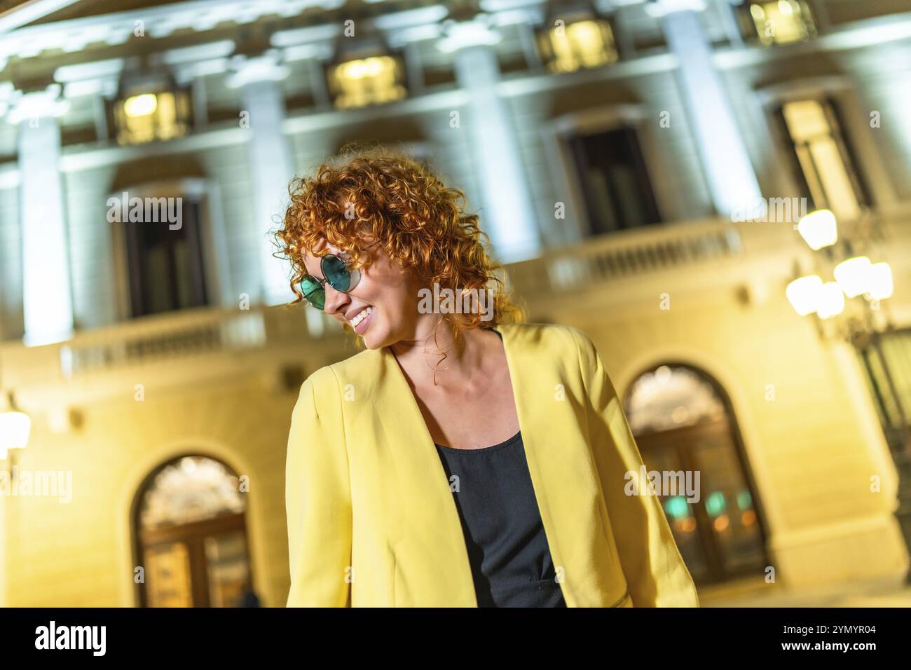 Vorderansicht einer stilvollen Frau mit legerer gelber Jacke und Sonnenbrille, die nachts in die Stadt schaut Stockfoto