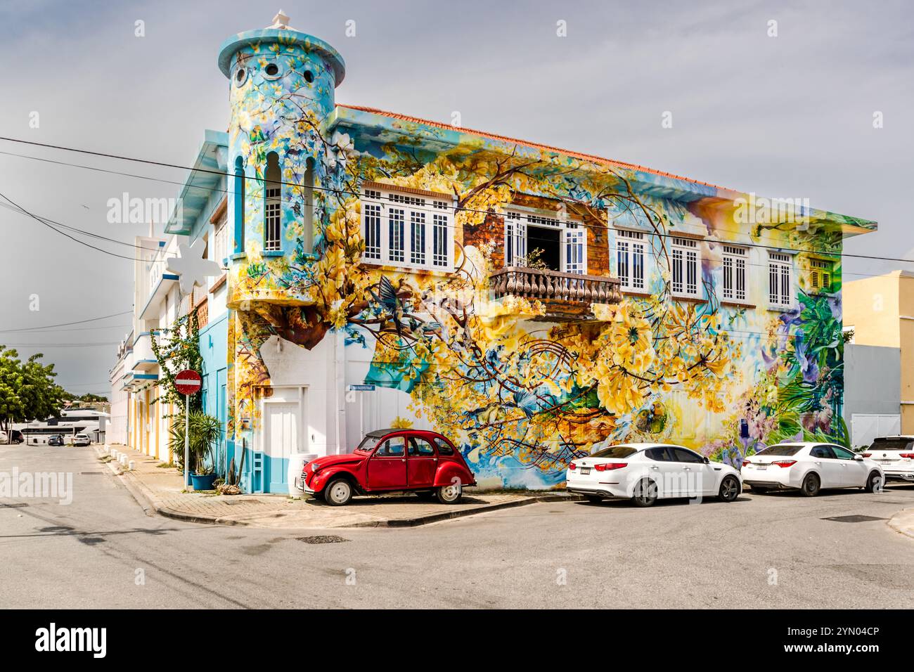 Das Wandgemälde „3 o'Clock Romance“ des Künstlers Francis Sling bedeckt sein Haus in der Van Raderstraat im Bezirk Scharloo von Willemstad, Curaçao, vollständig. Gelb blühende Bäume und blaue Vögel schmücken die Fassade und den abgerundeten EckTurm des historischen Gebäudes. Laut Sling zeigt das Motiv die Begegnung zweier Vögel auf einem Ast und gehört zu den bekanntesten Wandgemälden im Bezirk. Es wurde im Rahmen der Initiative „Street Art Skalo“ geschaffen, die Scharloo durch Kunst wiederbelebt. Immer häufiger schmücken solche Wandgemälde Häuser in Willemstad und verleihen jedem Viertel seine eigene Identität. Stockfoto