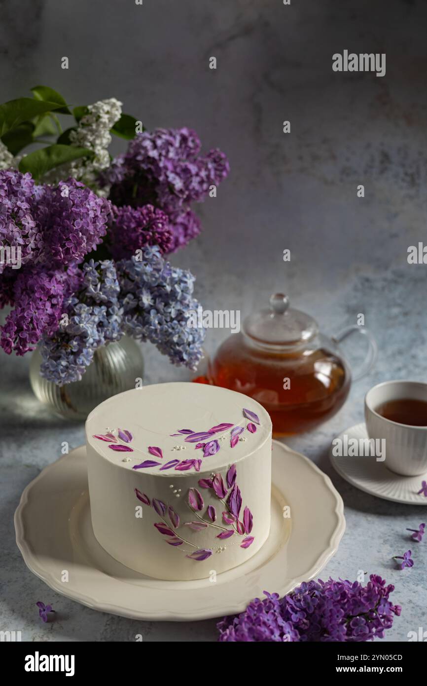 Eleganter weißer Kuchen mit lila Blumendekorationen auf einer Keramikplatte, umgeben von blühenden Fliedern, einer Teekanne und einer Tasse Tee Stockfoto