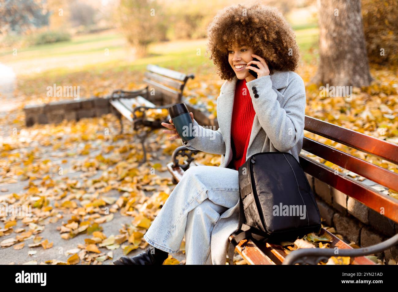 Gemütlich auf einer Bank sitzend, spricht eine fröhliche junge Frau mit lockigen Haaren auf ihrem Telefon, während sie eine Tasse Kaffee genießt, während ich im herbstlichen Herbstlaub i Stockfoto