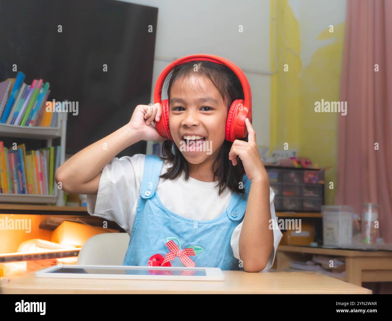 Niedliches asiatisches Mädchen, das Kopfhörer trägt, Musik hört und fröhlich in ihrem Klassenzimmer zu Hause tanzt. Stockfoto