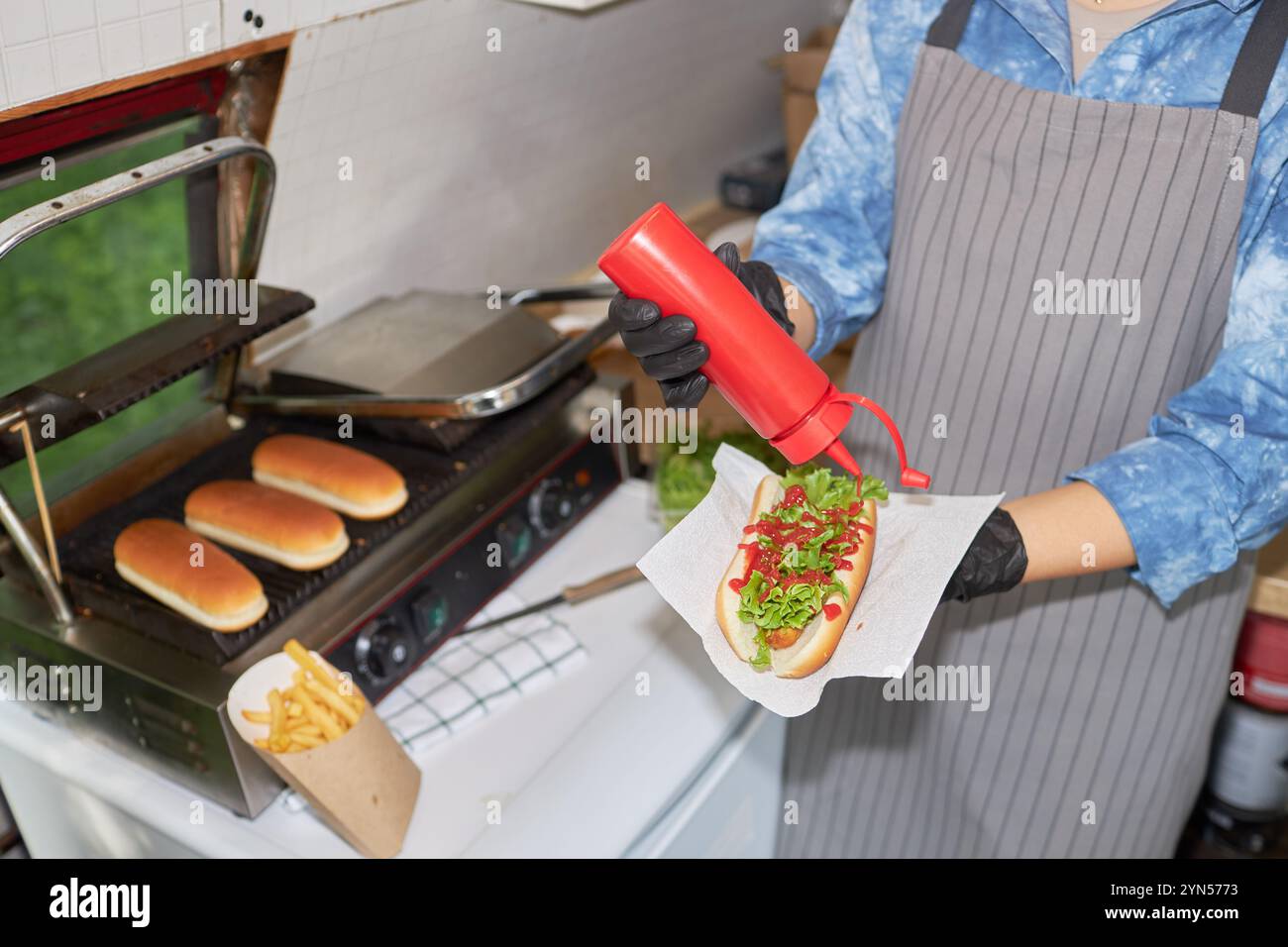 Hochwinkelaufnahme einer nicht erkennbaren Küchenchefin, die Ketchup in Hot Dog hinzufügt, während sie neben dem Grill mit Brötchen steht Stockfoto