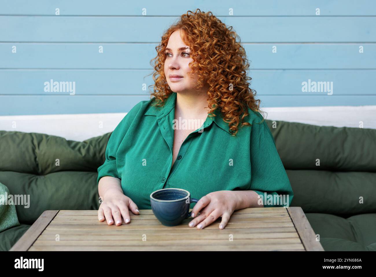 Schöne große Frau mit rotem Haar, die eine Tasse Kaffee genießt. Kurviges weibliches Model sitzt am Cafe-Tisch in grünem Kleid gegen helles buntes blaues W Stockfoto