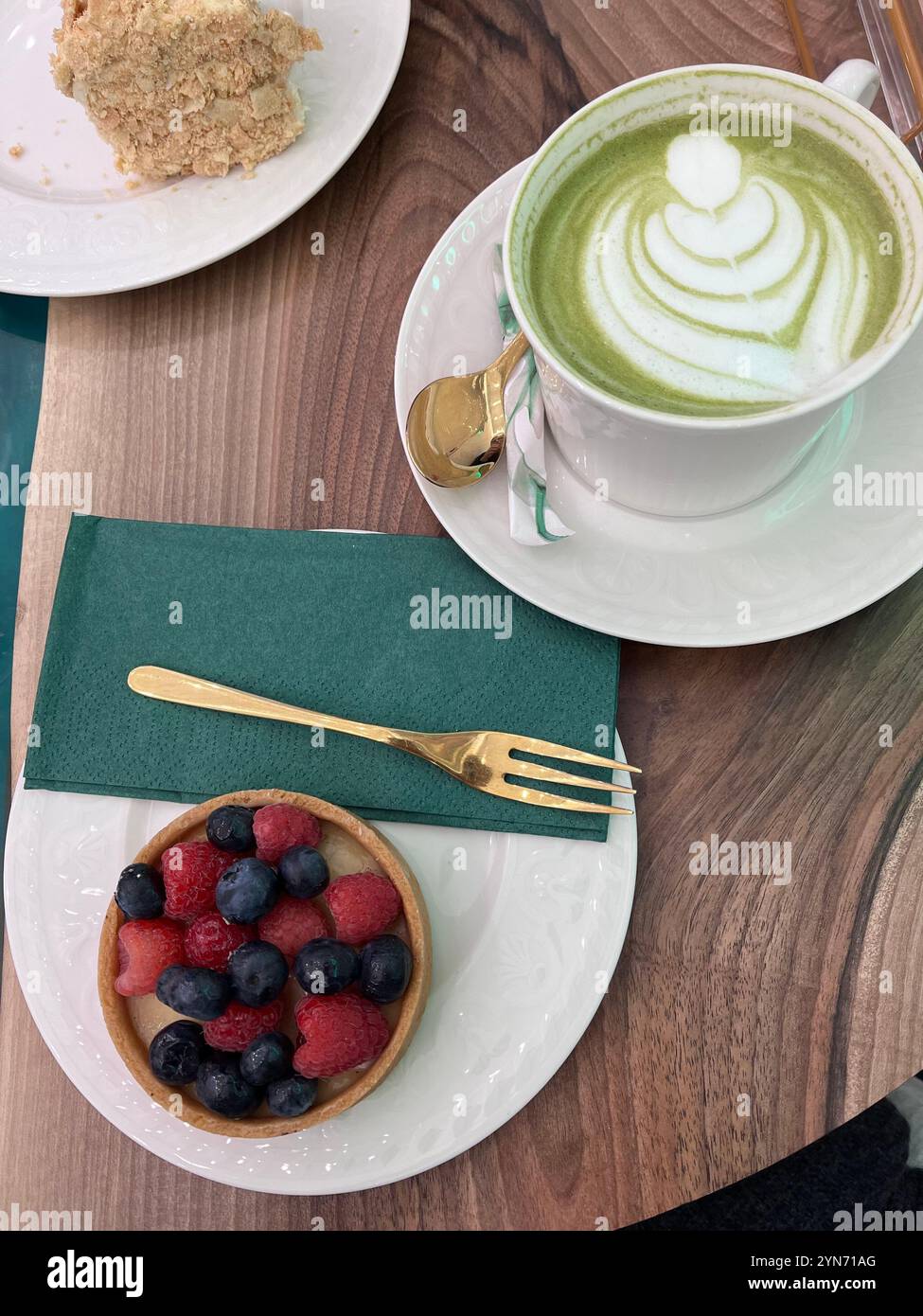 Ein Matcha Latte mit Herzmustern ruht neben einer farbenfrohen Obsttarte mit Himbeeren und Heidelbeeren. Die süßen Leckereien werden auf einem serviert Stockfoto