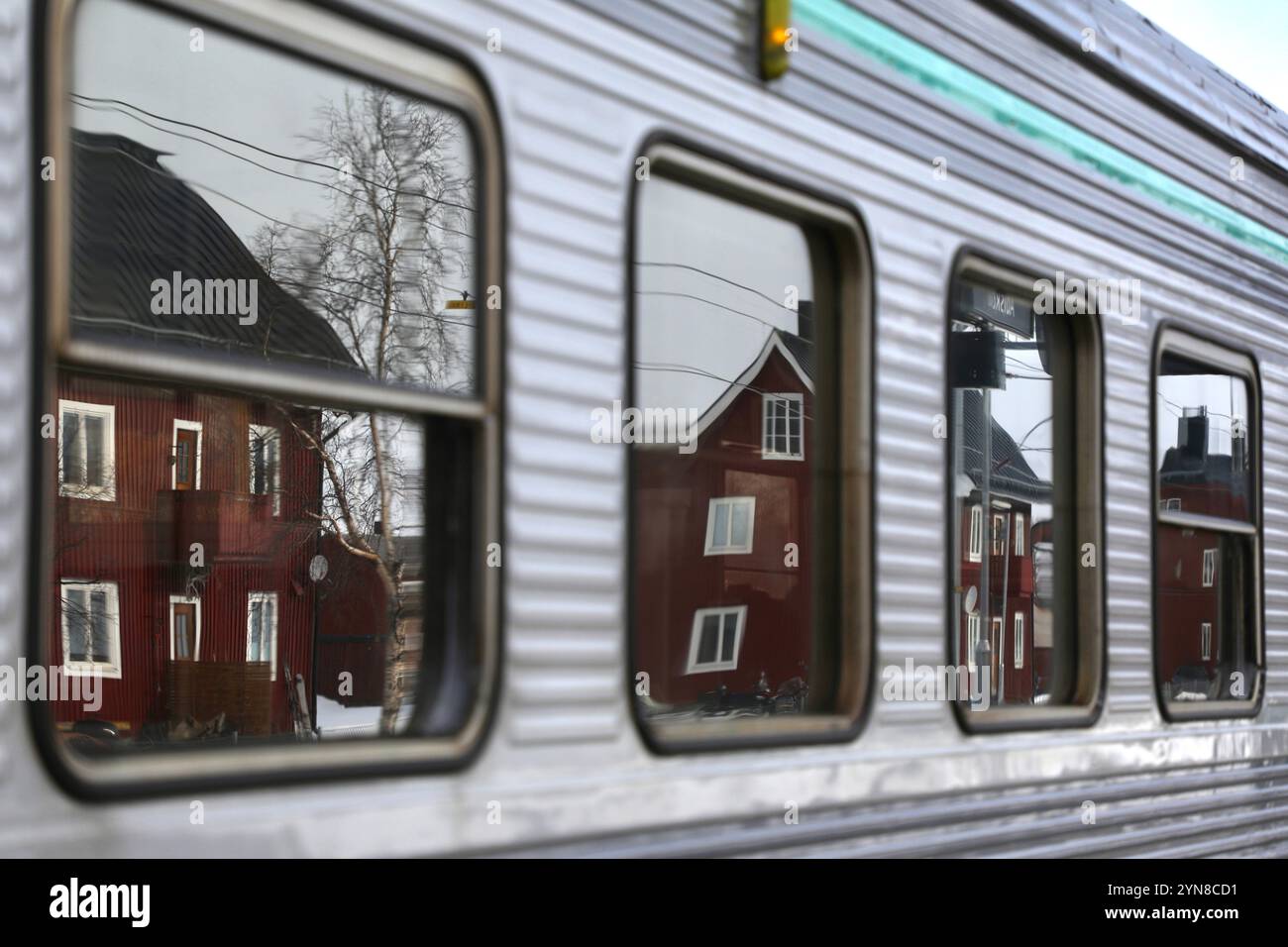 Die Fassaden des Dorfes Abisko spiegeln sich in den Fenstern eines Zuges wider, der an einem der Bahnhöfe Abiskos hält. Die Bahnstrecke wurde Anfang des 20. Jahrhunderts gebaut, als das erste Eisenerz in Kiruna, Nordschweden, abgebaut wurde. Es war die erste Eisenbahnstrecke jenseits des Polarkreises. Ursprünglich wurde es für den Transport schwedischen Erzes zum norwegischen Hafen Narvik genutzt. Bis in die 1980er Jahre gab es keine Straße zwischen den beiden Städten. Die Strecke ist heute noch mit Eisen bestückt, hat sich aber auch für den Personenverkehr geöffnet. Sie ist nach Murma die zweitnördlichste Personenlinie der Welt Stockfoto