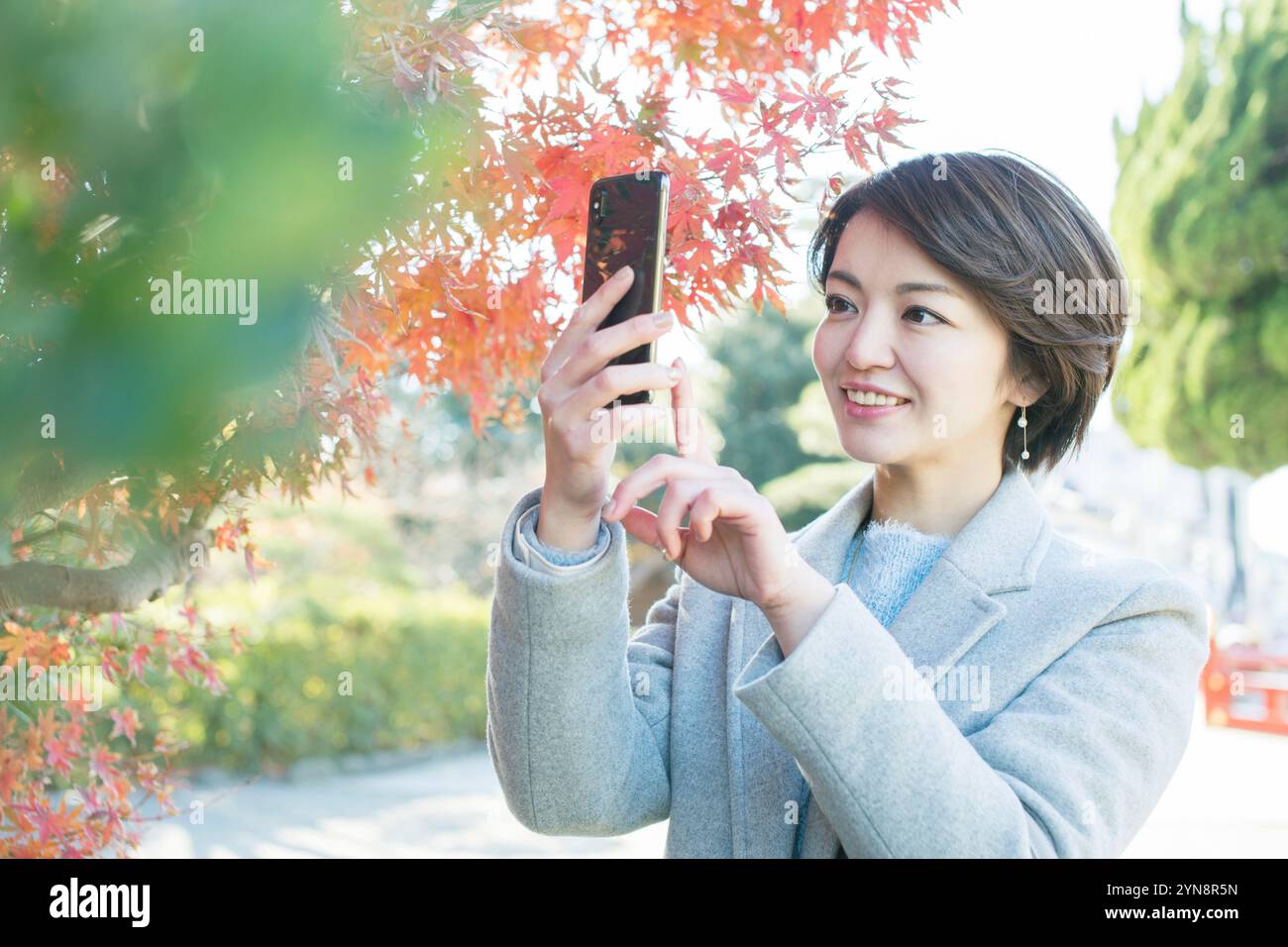 Lächelnde Frau in ihren 30ern, die ein Foto von Herbstlaub mit ihrem Handy macht. Stockfoto