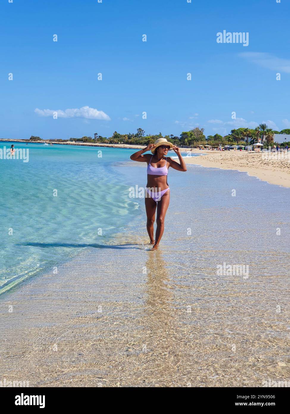 Eine Frau spaziert anmutig durch das flache Wasser an einem sonnigen Strand und genießt die Kühle des Ozeans, während sie einen stilvollen Badeanzug und einen Hut mit breiter Krempe trägt. San Vito Lo Capo Sizilien Italien Stockfoto