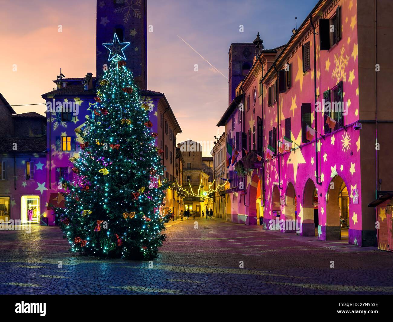 Ein Weihnachtsbaum mit funkelnden Lichtern im Zentrum des historischen italienischen Stadtplatzes, während farbenfrohe Projektionen die umliegenden Gebäude beleuchten. Stockfoto