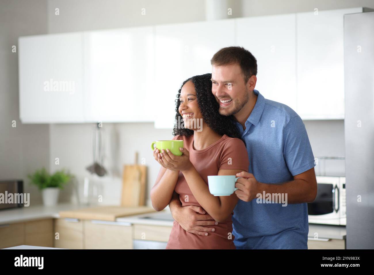 Glückliches, interrassisches Paar trinkt und sieht daheim in der Küche weg Stockfoto