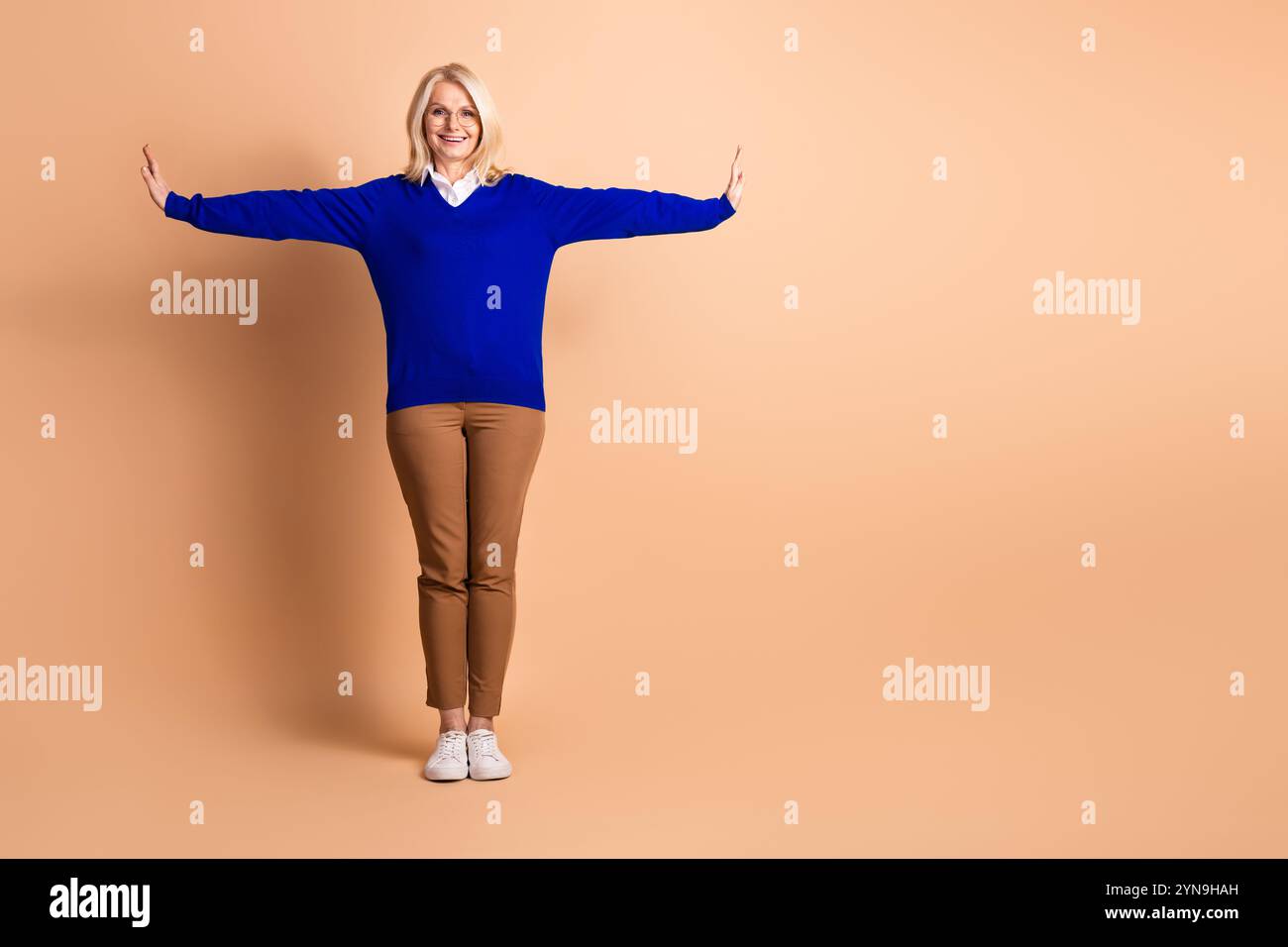 Elegante ältere Frau in blauem Pullover und legeren Hosen mit ausgestreckten Armen vor beigefarbenem Hintergrund Stockfoto