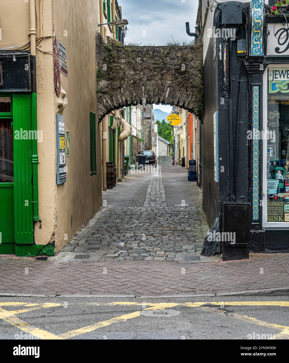 Pulsierende urbane Straße in Killarney, Irland, gesäumt von bunten Geschäften und Flaggen. Die Menschen schlendern durch die geschäftige Szene unter bewölktem Himmel, umgeben von Hist Stockfoto