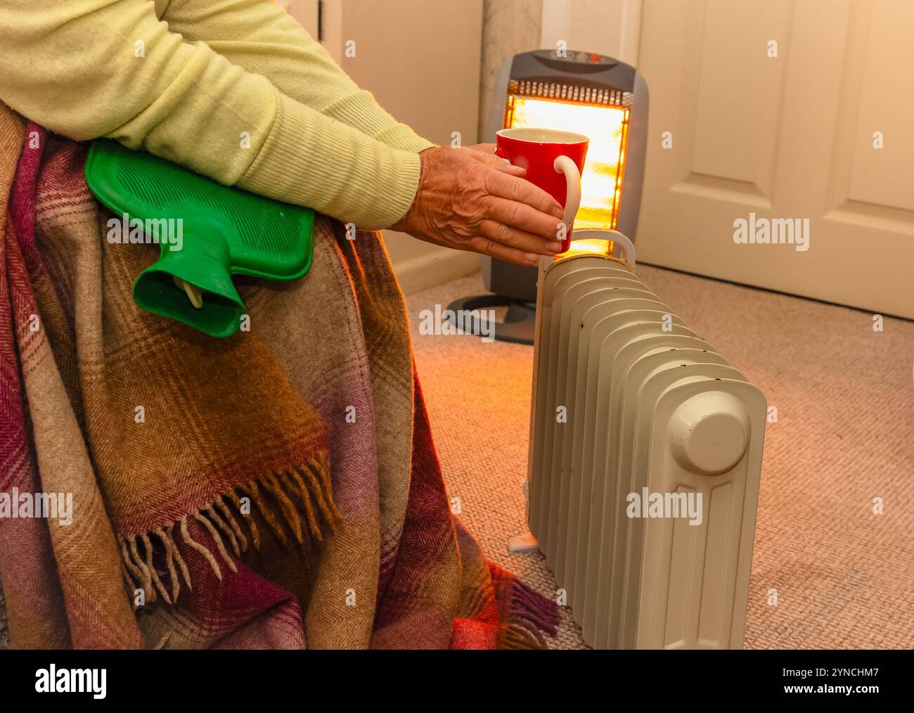 Rentner, der versucht, sich in der Nähe eines ölgefüllten Heizkörpers und eines Halogenheizgeräts mit einer Wärmflasche, einer Decke und einem warmen Getränk warm zu halten. 2024 wurde die Labour Gover Stockfoto