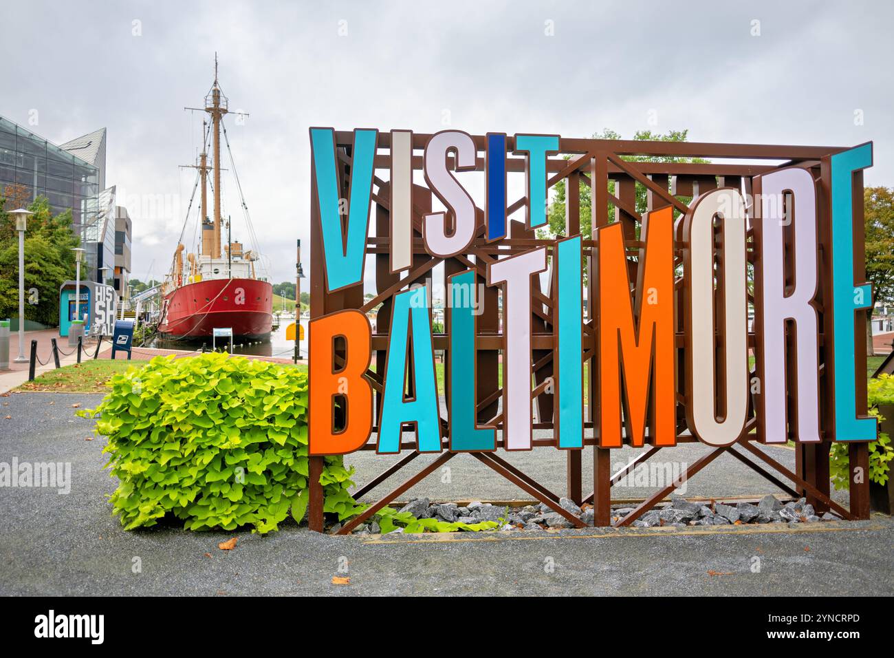 Besuchen Sie Baltimore Schild Inner Harbor Baltimore // BALTIMORE, Maryland - Ein Tourismusschild „Visit Baltimore“ steht am Inner Harbor deutlich sichtbar mit dem unverwechselbaren roten Rumpf des USCG Lightship Chesapeake im Hintergrund. Die Lightship Chesapeake, heute ein Museumsschiff, diente von 1930 bis 1971 als schwimmender Leuchtturm für die United States Coast Guard und leitete den Seeverkehr in der Chesapeake Bay. Das historische Schiff liegt dauerhaft im Inner Harbor von Baltimore als Teil der Historic Ships in Baltimore Collection, die das maritime Erbe der Chesapeake Bay regi bewahrt Stockfoto