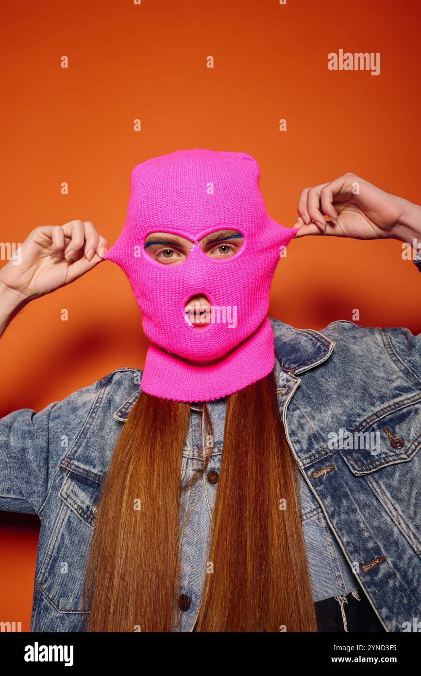 Ein junger Mann mit langen roten Haaren trägt eine pinkfarbene Maske vor einem lebendigen Hintergrund. Stockfoto