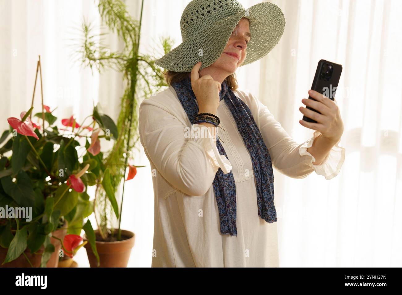 Eine Frau mit einem großen Strohhut und einem Schal macht mit ihrem Smartphone ein Selfie in einem hellen Raum voller Pflanzen. Sonnenlicht filtert durch sie Stockfoto