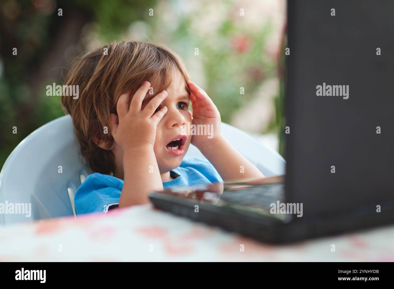 Ein kleines Kind sitzt an einem Tisch und greift überraschend oder ängstlich den Kopf, während es auf den Computerbildschirm blickt. Die Szene fängt einen intensiven Moment der Emoti ein Stockfoto