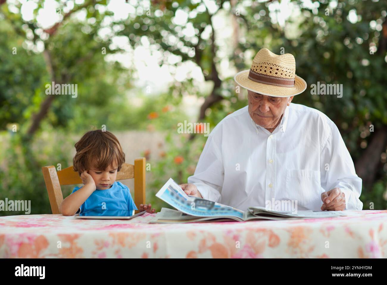 Eine berührende Szene, die den Kontrast zwischen Generationen zeigt. Ein Großvater liest eine Zeitung, während sein kleiner Enkel sitzt Stockfoto