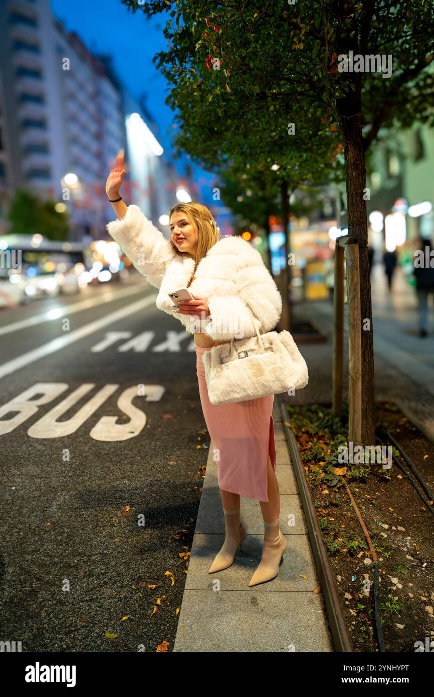 Modische Influencerin, die nachts ein Taxi in der Stadt ruft, ihr Smartphone und eine Handtasche in der Hand hält Stockfoto