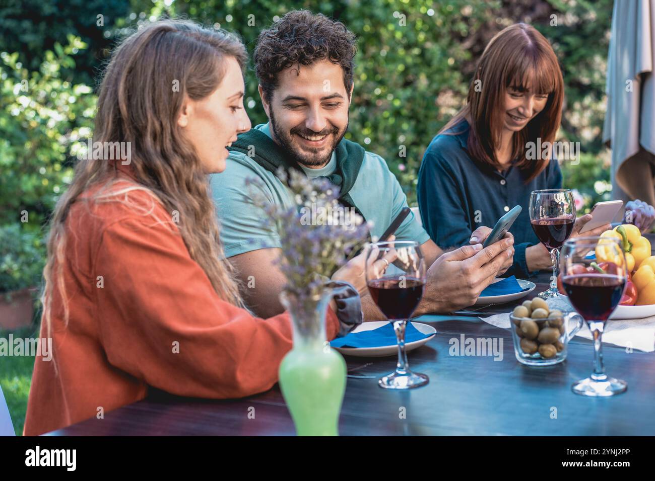 Moderne soziale Interaktion von Freunden mit Smartphones während eines zwanglosen Abendessens im Freien. Zeitgenössischer Lifestyle-Moment, der digitale Verbindung mit Re Stockfoto