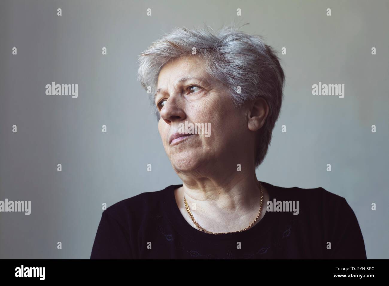 Eine Frau mit grauem Haar und einem kontemplativen Ausdruck schaut zur Seite. Weiches Tageslicht hebt ihre Eigenschaften hervor und schafft eine ruhige Atmosphäre. Sie Stockfoto