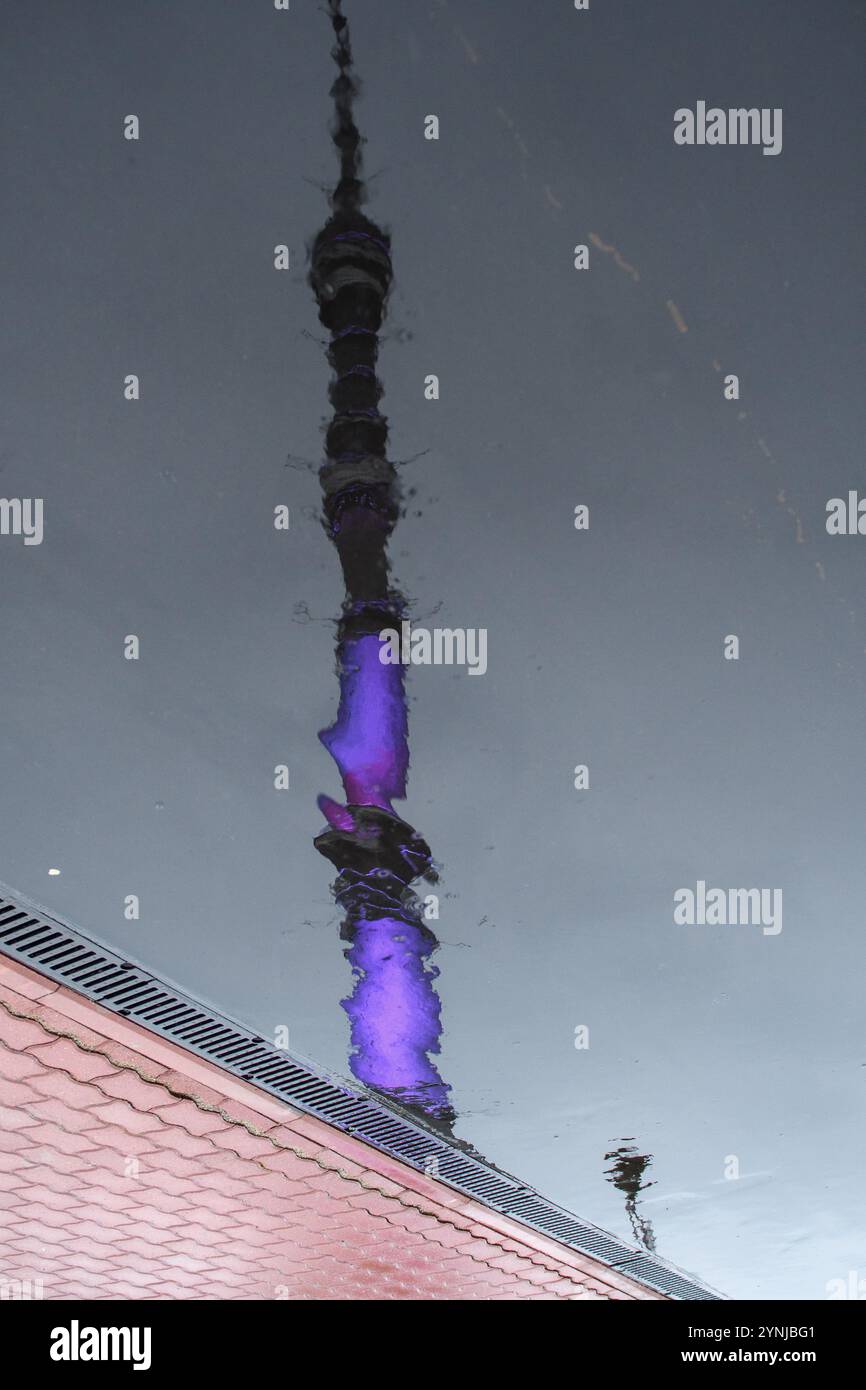 Die beleuchtete Reflexion des Ostankino-Turms in einer Regenpfütze erzeugt einen beeindruckenden visuellen Kontrast zum nassen Gehweg. Stockfoto