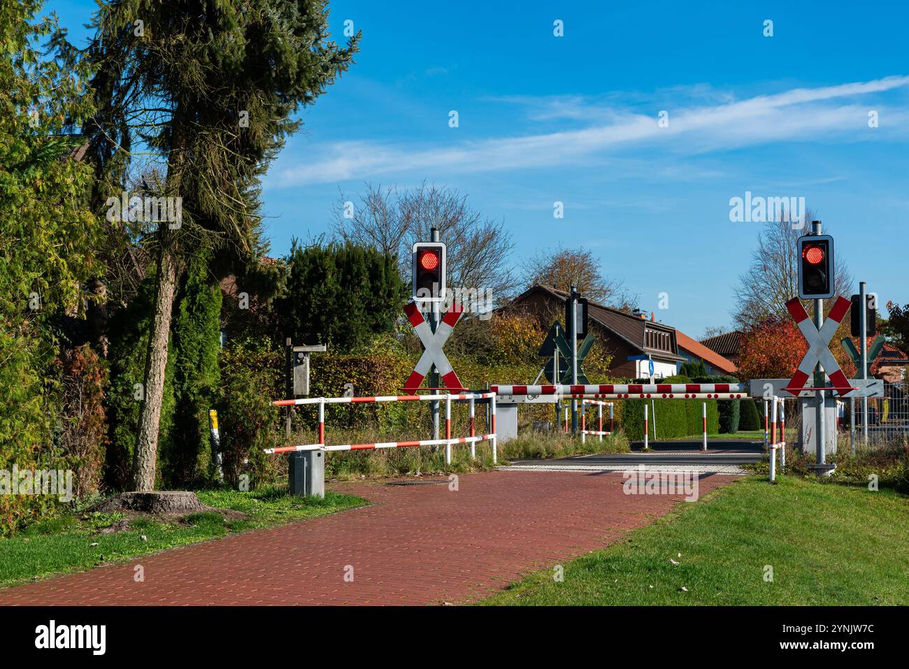 Leuchtend rote Warnleuchten beleuchten die geschlossenen Bahnübergänge in einer friedlichen ländlichen Umgebung mit Bäumen und Häusern vor einem klaren blauen Himmel. Stockfoto