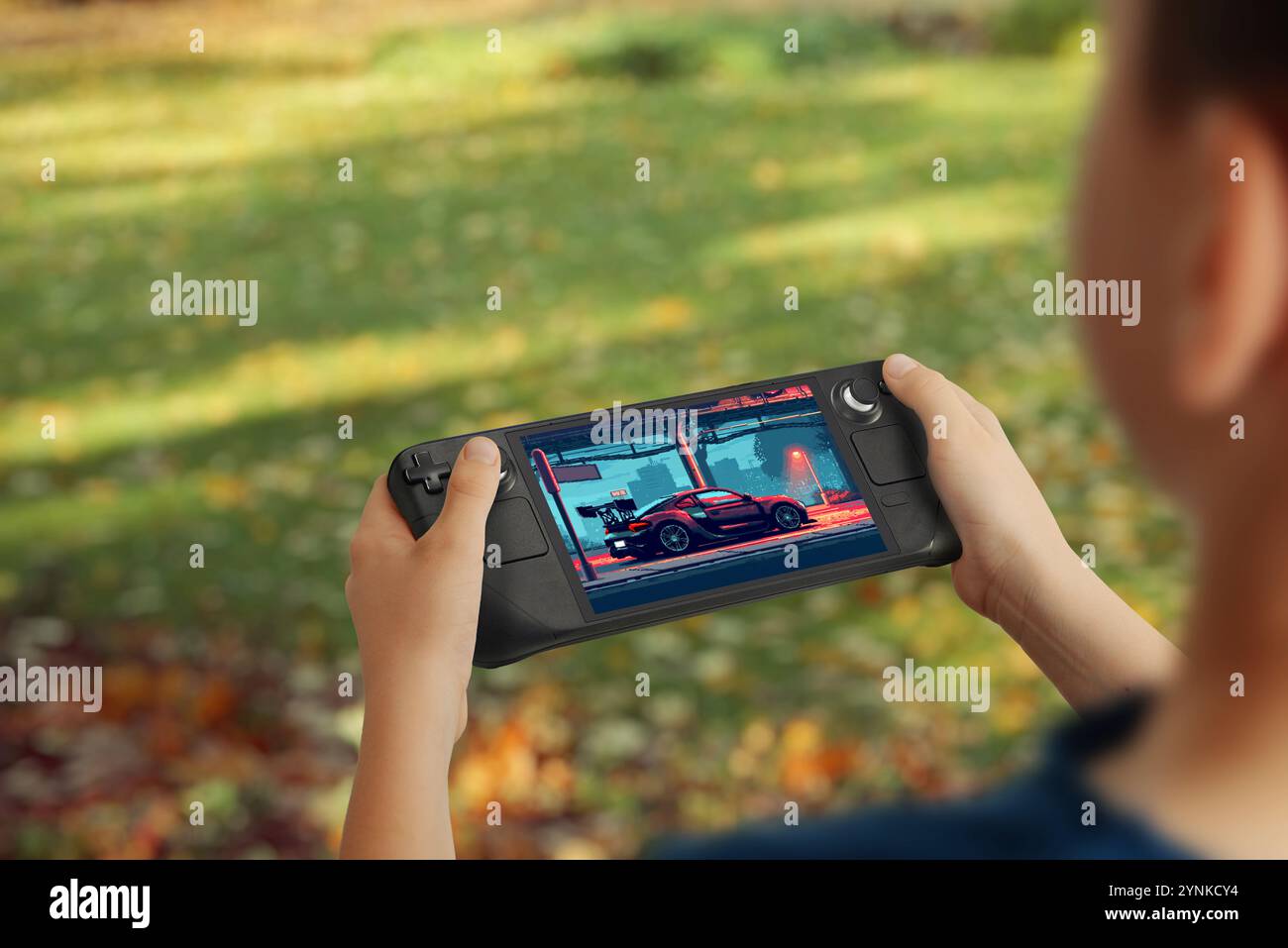 Junge, der eine Handheld-Spielkonsole hält, ein Fahrspiel mit einem Auto auf dem Bildschirm spielt, Park-Einstellung im Freien, Hervorhebung von tragbaren und interaktiven Spielen Stockfoto