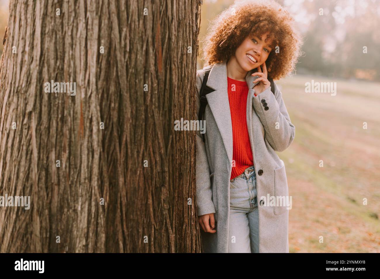 Eine lächelnde junge Frau mit lockigem Haar steht vor einem Baum und trägt ein gemütliches Outfit. Sie liebt die Schönheit des Herbstes in einem ruhigen Park voller Colo Stockfoto