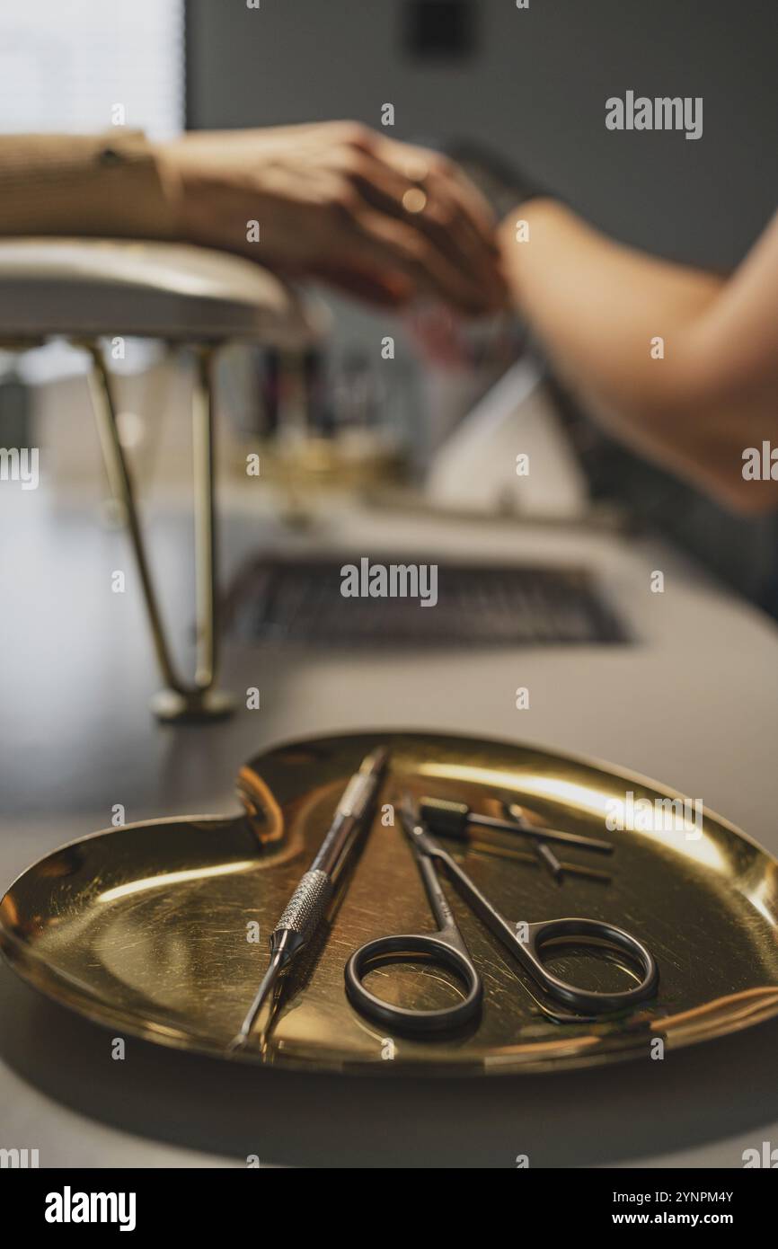 Werkzeuge für die Herstellung von Nägeln in Schönheitssalon, Schere, Cutter auf einem goldenen Herzständer. Kunde mit Kosmetologe im Hintergrund Stockfoto