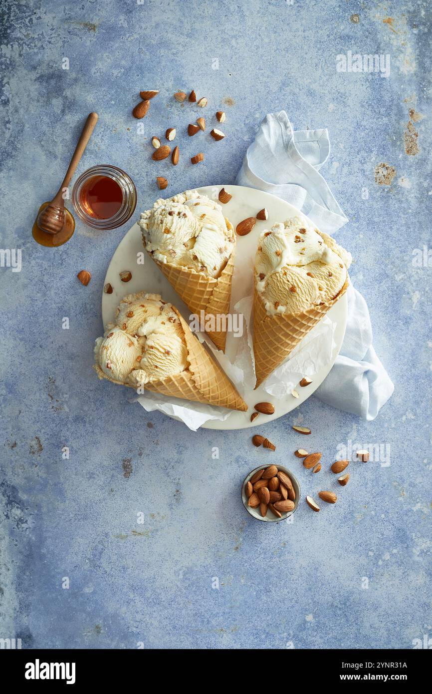 Mandel-Honig-Eis in drei knusprigen Eiskegeln zubereitet, auf einer weißen Platte angeordnet, mit Mandeln und Honig auf blauem Hintergrund dekoriert Stockfoto