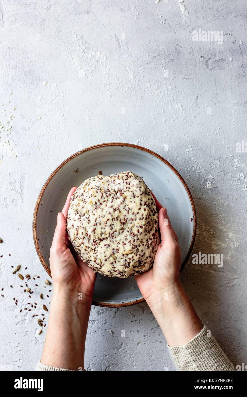 Eine Person, die eine runde Kugel Teig mit Samen in einer Keramikschale auf einer strukturierten Oberfläche hält. Stockfoto