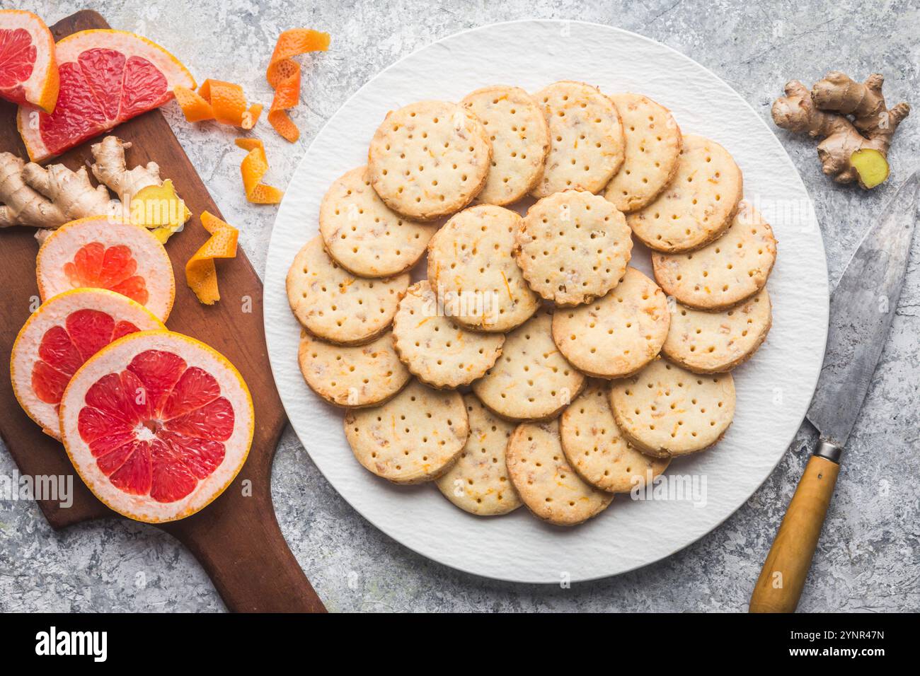 Ein weißer Teller, hoch gestapelt mit hausgemachten Ingwer- und Grapefruit-Shortbread-Keksen auf einem benutzerdefinierten grauen Hintergrund. Ein hölzernes Schneidebrett hält Scheiben von fr Stockfoto