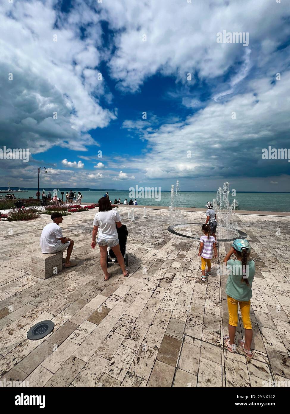 Tag am Wasser, während Familien entlang der Promenade spazieren und Kinder in der Nähe von Springbrunnen spielen. Panoramablick auf das Meer, ergänzt durch dramatische Wolken o Stockfoto