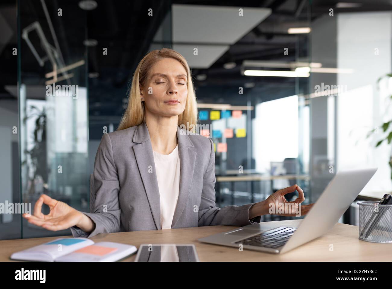 Reife Geschäftsfrau, die sich in der Achtsamkeitsmeditation während der Büropause engagiert. Sie sitzt ruhig vor dem Laptop und sucht nach innerer Ruhe inmitten von Stress am Arbeitsplatz und täglichen Herausforderungen. Stockfoto